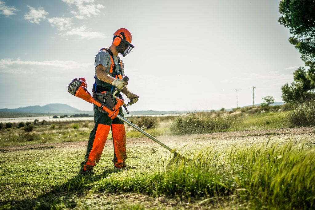 Backlit side view of a man wearing protective gear removing grass with a trimmer outside a house on a sunny spring afternoon. Great detail of the grass splashing through the brush.